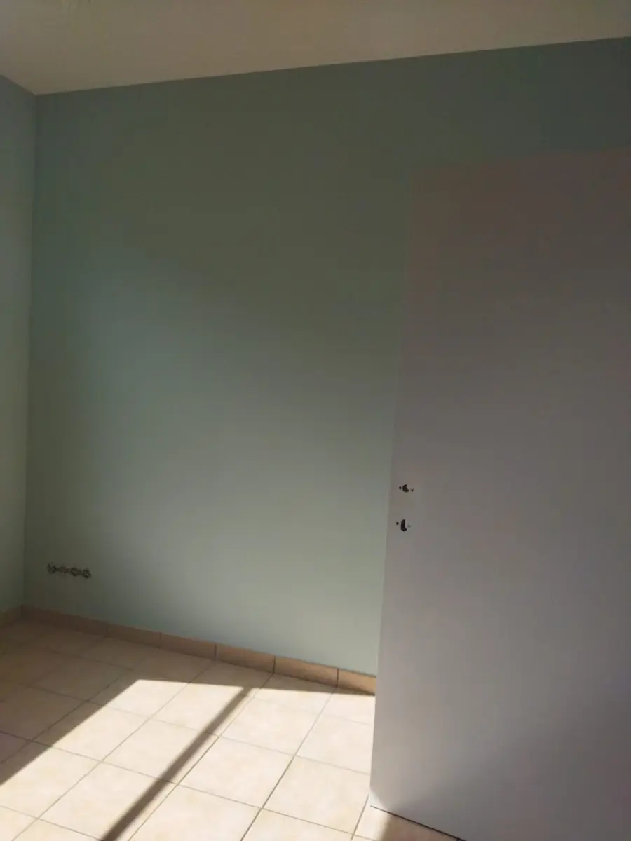 Kamer met een grijsgroen geschilderde muur, een witte deur en zonlicht op de tegelvloer